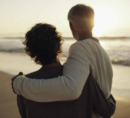 Två personer står på en strand och ser ut över havet i solnedgången.