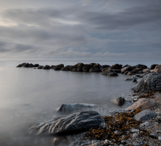 En stenig strand vid havet med molnig himmel i bakgrunden.