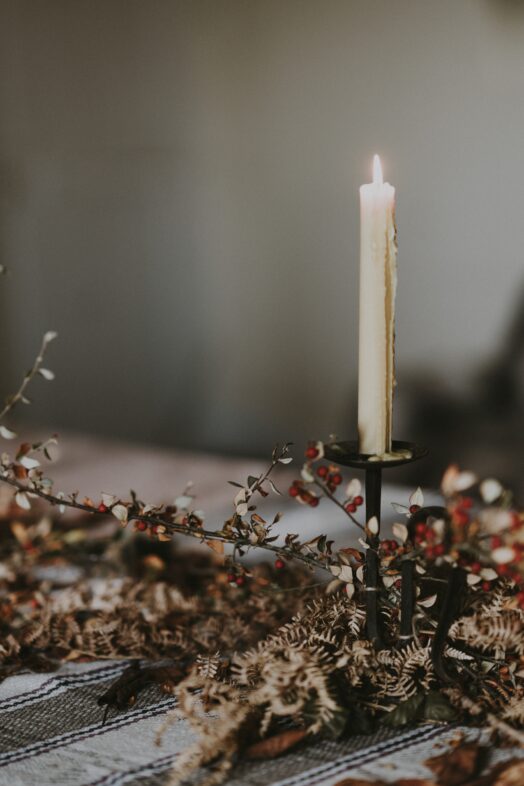 Ett tänt stearinljus i en mörk ljusstake omgiven av torkade blommor på ett bord.