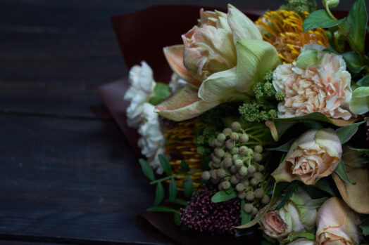 Närbild av en bukett med olika blommor och gröna blad på ett mörkt bord.